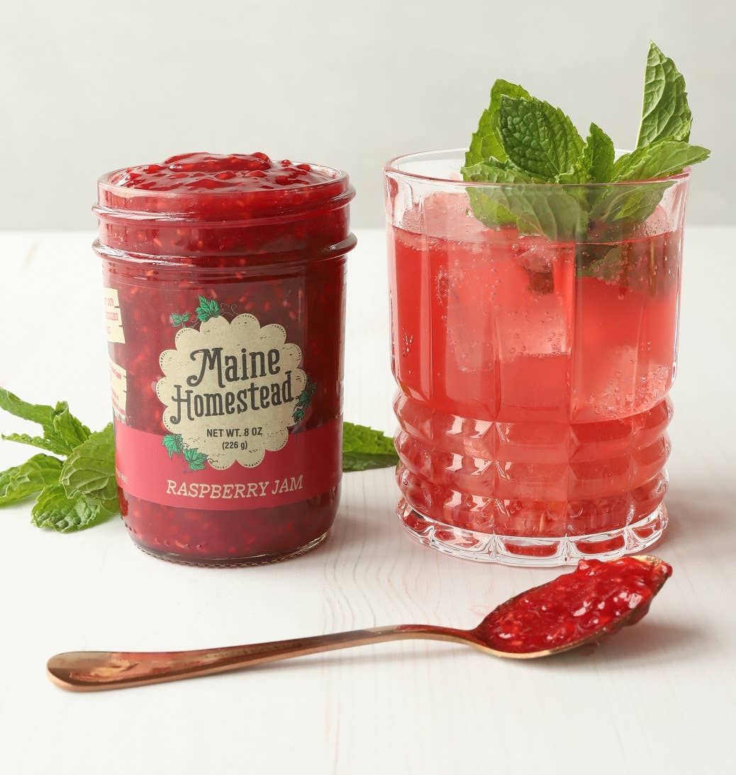 Jar of Maine Homestead raspberry jam next to a glass of red drink with mint leaves on a white background