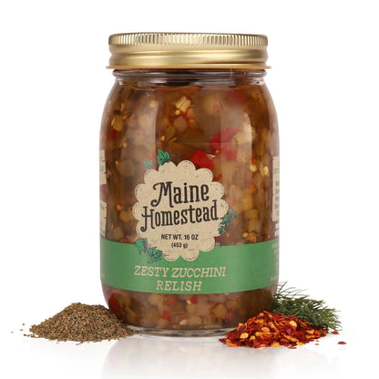 A jar of Maine Homestead Zesty Zucchini Relish with a gold metal lid and a green and cream label, shown on a white background with piles of seasoning and herbs at the base.