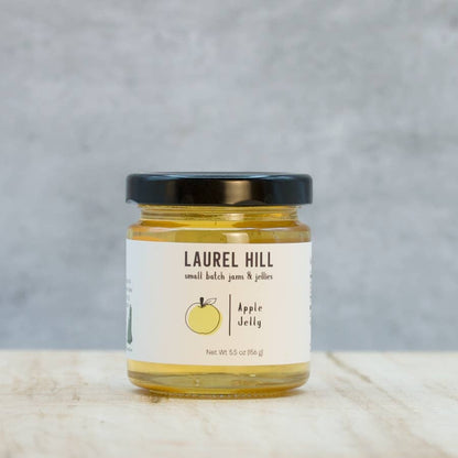 A jar of Laurel Hill Apple Jelly on a light wood surface with a textured gray background. The clear glass jar shows the golden-yellow jelly inside, topped with a black lid and featuring a white label with a yellow apple illustration.