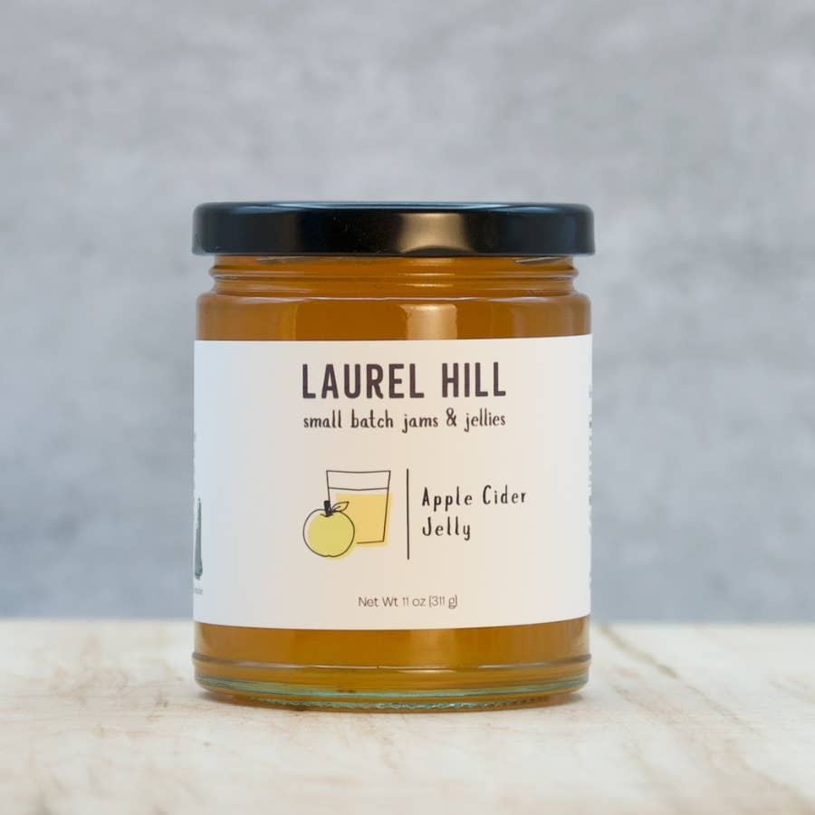 A jar of Laurel Hill Apple Cider Jelly on a light wooden surface with a textured gray background. The glass jar contains amber-colored jelly and has a cream-colored label with a black lid.