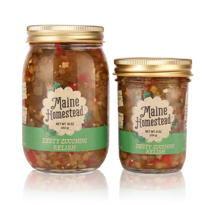 Two glass jars of Maine Homestead Zesty Zucchini Relish with gold lids, one 16 oz and one 8 oz, on a white background with reflections.
