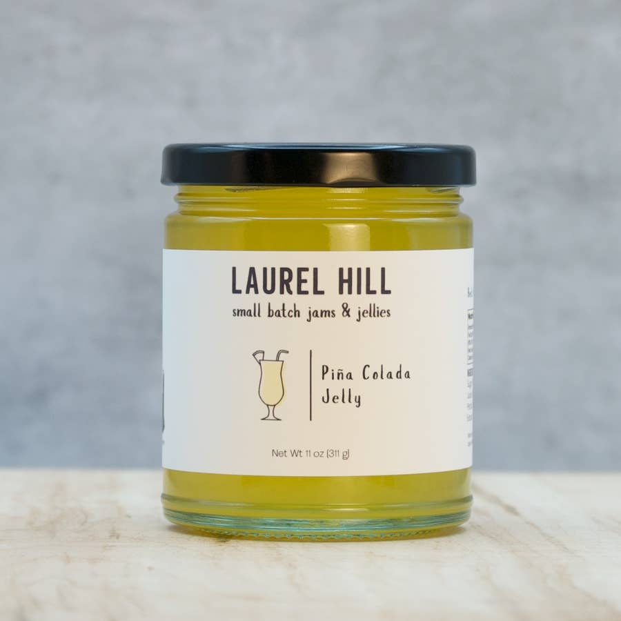 A jar of Laurel Hill Piña Colada Jelly on a light marble surface with a textured gray background. The clear glass jar shows the yellow-green jelly inside, has a black lid, and a white label with black text and a cocktail glass illustration.