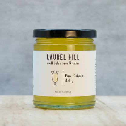 A jar of Laurel Hill Piña Colada Jelly on a light marble surface with a textured gray background. The clear glass jar shows the yellow-green jelly inside, has a black lid, and a white label with black text and a cocktail glass illustration.