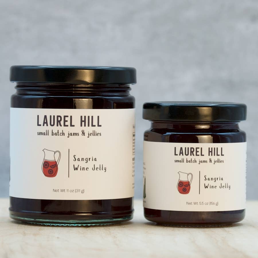 Two glass jars of Laurel Hill Sangria Wine Jelly on a light stone surface against a soft gray background. The larger jar on the left and the smaller jar on the right both have black lids and white labels with black text and a small red pitcher illustration.
