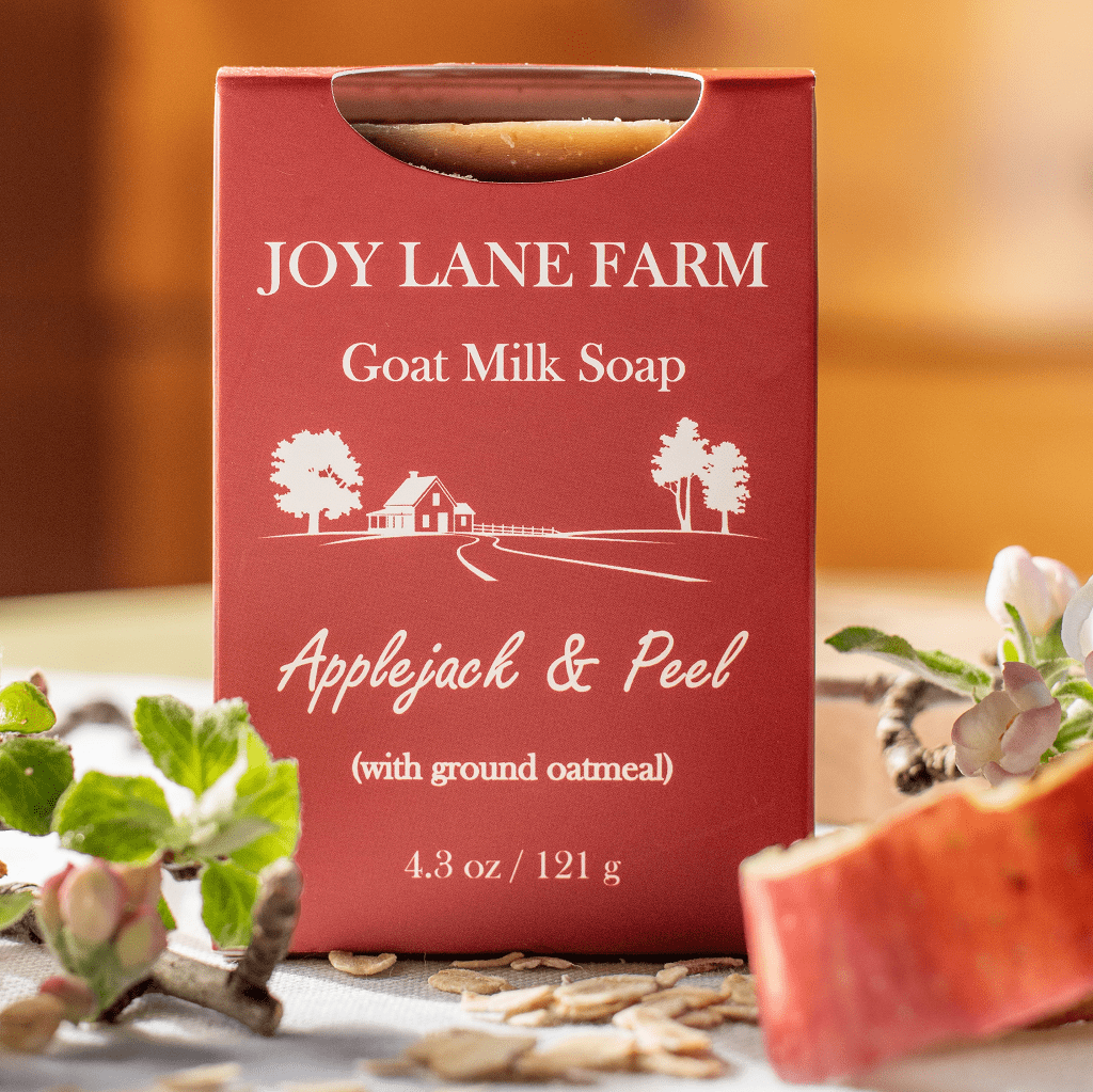 A red box of Joy Lane Farm Goat Milk Soap, Applejack & Peel, styled on a table with apple slices, oats, and flowers.