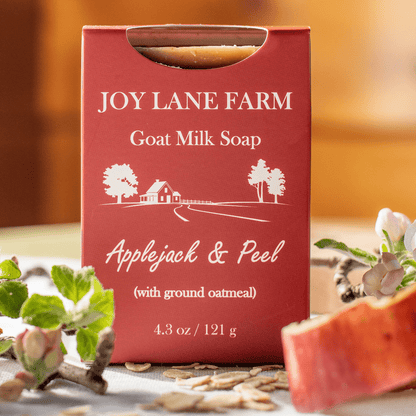 A red box of Joy Lane Farm Goat Milk Soap, Applejack & Peel, styled on a table with apple slices, oats, and flowers.