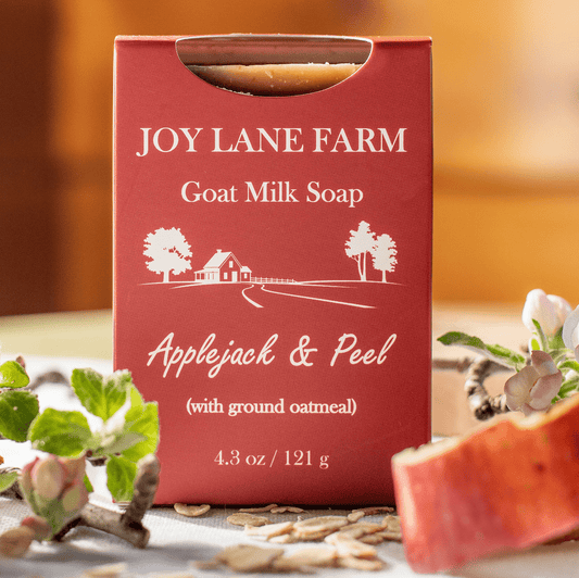 A red box of Joy Lane Farm Goat Milk Soap, Applejack & Peel, styled on a table with apple slices, oats, and flowers.