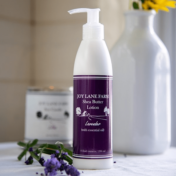 Joy Lane Farm Shea Butter Lotion in a white pump bottle with a purple label, styled on a table with lavender flowers, a white vase, and a candle in the background.