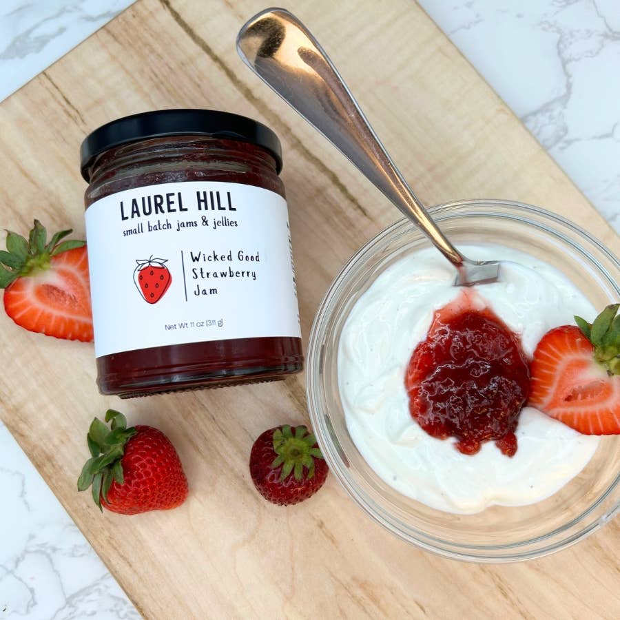 Jar of Laurel Hill Wicked Good Strawberry Jam on a wooden board with a spoon, bowl of yogurt, and strawberries.