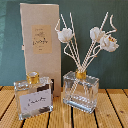 Clear perfume bottle with gold cap and white floral diffuser on a green and wooden surface.